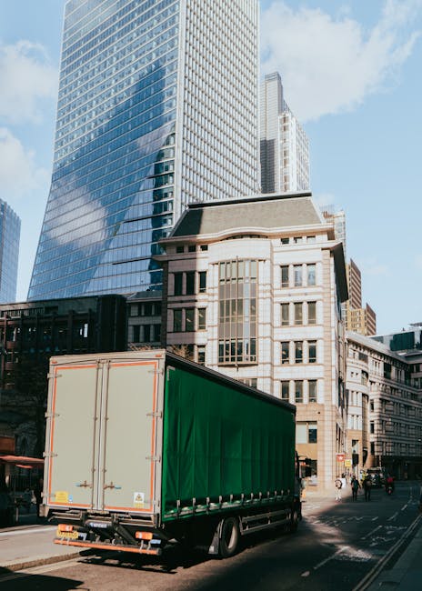 A large green and beige box truck parked on a city street during daylight hours, surrounded by tall modern office buildings and older architectural structures. The truck is positioned near the sidewal