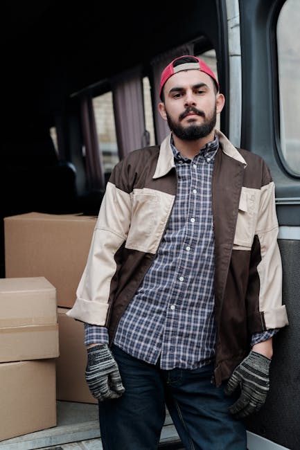 A young man with a beard, wearing a pink cap, plaid shirt, and a beige and brown work jacket, stands inside a moving van surrounded by cardboard boxes. The boxes are stacked on the van's floor and are